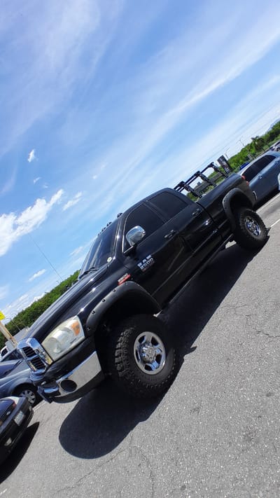 A black Dodge Ram 2500 pickup truck is parked on an asphalt surface, likely a parking lot. The truck is angled, with its front passenger side facing the camera. The truck has chrome rims, fender flares, and a rack system over the bed. The background shows a clear blue sky with some wispy clouds, green trees, and other vehicles parked in the distance. The scene appears to be in a sunny outdoor environment, with the shadow of the truck cast on the ground.