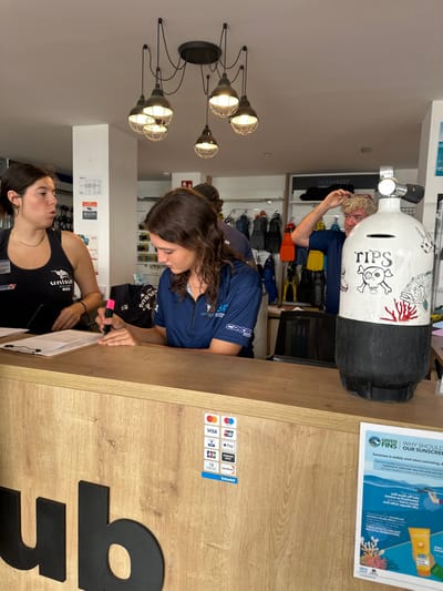 The image portrays the interior of a dive center in L'Estartit, Spain, confirmed by "L'ESTARTIT" visible on a person's shirt. The central focus is a light-colored wooden reception counter.

Three individuals are present. On the left, a young adult female with dark hair, wearing a black tank top with "UNISUB" and "Cressi" logos, stands behind the counter, looking right with her mouth slightly open. In the center, a second young adult female with long, wavy dark hair, wearing a dark blue polo shirt bearing "L'ESTARTIT" and "CRESSI" branding, sits at the counter, actively writing on a document placed on a clipboard with a pink highlighter. Partially obscured behind the central female, the back of another person in a dark shirt is visible. To the right of the central female, a man with light, curly hair and a blue t-shirt is visible in the background, raising his left hand towards his head.

On the counter's surface, a decorative white and black scuba tank serves as a "TIPS" jar, featuring a skull and crossbones, red coral, and a fish illustration. Payment method decals for Visa, Mastercard, Apple Pay, Google Pay, and Sabadell are affixed to the counter's front, where large black letters "ub" are partially visible. A black monitor or laptop screen is visible behind the tip jar.

The background reveals shelving units displaying various diving equipment, including fins, wetsuits, and masks. An "OCEANREEF" sign is visible above the shelves. Overhead, a black spider-style chandelier with seven caged industrial-style light fixtures illuminates the space. On the far right, a "GREEN FINS" poster advises on eco-friendly sunscreen.