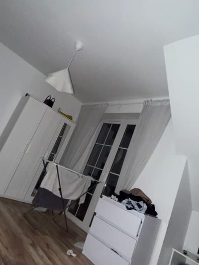 The image shows an indoor scene, likely a bedroom, in Falenty Nowe, Poland. The room is sparsely furnished and appears to be in a state of disarray. A large white wardrobe stands against the left wall. In the center of the room, a drying rack is set up with laundry draped over it. Behind the drying rack, a set of French doors with multiple panes leads outside. White sheer curtains partially obscure the doors. On the right side of the room, a chest of drawers is visible, with clothes piled on top. A ceiling light fixture hangs from the white ceiling. The floor is made of wood. The overall impression is one of a functional but untidy living space.