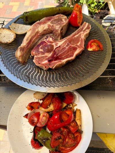Two plates of food are arranged on a grill. On the left, a metal grill pan holds two raw lamb chops, a whole green chili pepper, slices of white bread, and two tomato wedges. The lamb chops are a deep pink with streaks of white fat. The grill pan has a decorative pattern of wavy lines. On the right, a white plate is filled with grilled vegetables, including red bell pepper chunks, tomato halves, and pieces of what appear to be mushrooms. The vegetables show signs of charring from the grill. The scene is outdoors, likely during the daytime, given the bright lighting and the presence of greenery in the background. The setting appears to be a backyard or patio with a grill. There are no people visible in the image.