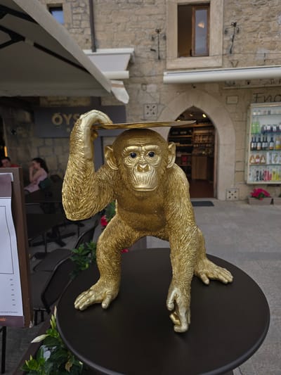 A golden monkey statue is posed in a crouching position on a dark round table, its left hand balanced on its head holding a flat, golden plate. The statue is positioned outdoors in front of a building with stone walls and an arched entryway. Behind the statue, an awning extends over a seating area with tables and chairs. To the right of the entryway, a display case filled with bottles is visible. The ground appears to be paved. The statue is the primary subject, with the background providing context of an outdoor dining or shopping area.