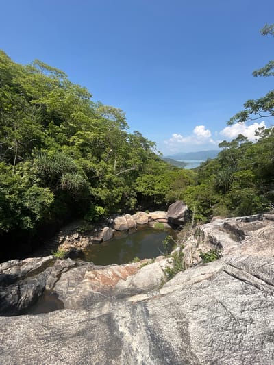 The image depicts a natural landscape scene in Hoa Thang Ward, Vietnam. In the foreground, rough, gray granite rocks form a natural ledge overlooking a small, dark pool of water. The pool is surrounded by more rocks, some of which have reddish-brown mineral deposits. Dense, lush green foliage and trees cover the slopes of the surrounding hills. In the middle ground, the landscape opens up to reveal a body of water, possibly a bay or a large river, with distant hills and mountains visible on the horizon. The sky is a clear, bright blue with a few scattered white clouds. The overall impression is one of a serene, natural environment.