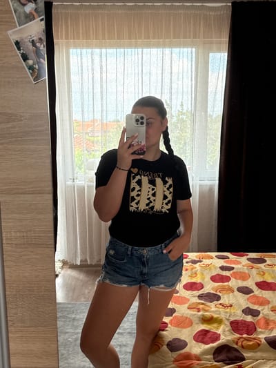 A young woman with dark hair in braids is taking a mirror selfie in a bedroom. She is wearing a black t-shirt with a gold and beige graphic design and distressed denim shorts. She is holding a white smartphone in her right hand. To the left of the woman, a closet door with a wood grain texture is visible, and on it are three framed photographs. Behind her, a window is covered by sheer white curtains, and outside the window, rooftops and trees can be seen. To the right of the window, dark brown curtains are drawn. The woman is standing on a light gray rug, and a bed with a colorful, patterned comforter is partially visible on the right side of the image. The location is stated as Rogosh, Bulgaria.