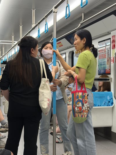 The image depicts the interior of a subway car in Tiaodeng, China. Three individuals are visible. On the left, a woman with dark hair pulled back stands with her back to the camera, wearing a black top and black pants, carrying a light-colored canvas tote bag. In the center, a woman wearing a light-colored surgical mask and a light blue shirt is looking towards the right. She is wearing light blue jeans and is holding onto a blue pole. To the right, a woman in a bright green t-shirt and light blue jeans is facing the woman in the mask. She is holding a colorful tote bag with cartoon characters and also gripping the blue pole. The environment shows handrails and advertising posters on the walls of the subway car.