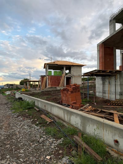 The image shows an ongoing construction site in what appears to be Cherni Vrah, Bulgaria, based on the buildings in the background. Several unfinished concrete and brick structures are visible, suggesting multiple units under construction. Piles of bricks are stacked near the structures. Construction materials, including wooden planks and support beams, are scattered throughout the site. A concrete retaining wall runs along the edge of the construction area, with grass and weeds growing along its base. The ground is covered in gravel and dirt. The sky is overcast with a mix of grey and blue clouds. In the distance, there are houses and power lines. A person wearing a high-visibility vest is briefly visible, further suggesting an active construction site.