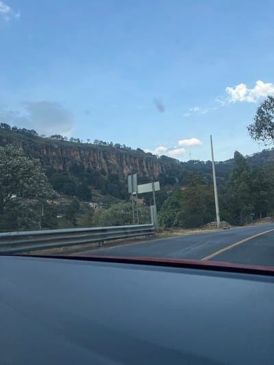 A view from a car window features a mountain or cliff face with visible rock layers and some vegetation. A road with a yellow stripe runs alongside the mountain, separated by a guardrail. A few signs are visible beside the road. The sky is blue with some clouds. The car's window and part of the car's frame are visible in the foreground, obscuring the lower right corner of the image.