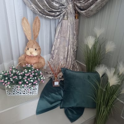 The image shows a still life arrangement in what appears to be an indoor setting, possibly a home or a shop display, in Briceni, Moldova. The arrangement features a stuffed straw bunny with long ears and pink accents, positioned next to a white basket filled with small pink and white faux flowers. Behind the bunny and flowers, a draped grey curtain with a damask pattern is visible, along with sheer white curtains. To the right of the bunny, there are tall faux plants with feathery white plumes, and a dark teal velvet pillow. In front of the pillow, a glass bottle of reed diffuser with wooden sticks is placed on a dark teal fabric. The background wall is paneled with light grey vertical slats. The floor is tiled with a beige marble pattern. There are no people or animals present in the image, and no discernible actions or interactions are taking place. The scene is static and decorative.