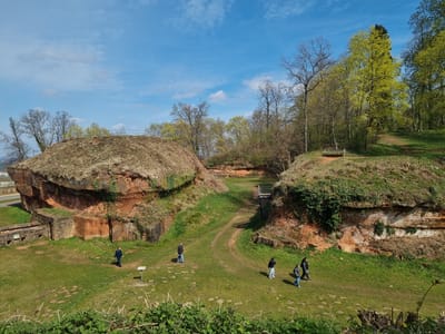 The image shows an outdoor scene at the Schlossberg Höhlen in Homburg, Germany. Several people are walking on a grassy area between large, red sandstone structures covered with vegetation. A small wooden staircase connects the ground level to the top of one of the structures. The sky is blue with scattered clouds, and trees are visible in the background, some with spring foliage. The overall setting appears to be a historical or archaeological site.
