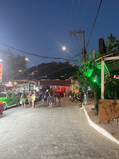 A bustling street scene at night showcases a lively gathering of people. A vintage green truck adorned with "Chopp Brahma" stands out. Above, electrical wires run along a pole, leading to a building with green lights. Further down the street, a group of people is visible, with a motorcycle parked nearby. A hill with buildings can be seen in the background. The sky is a dark blue gradient.