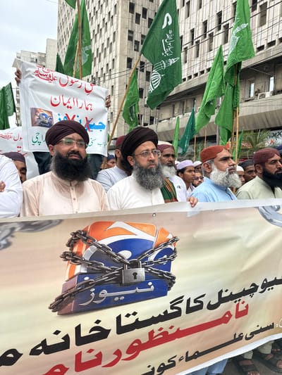 A group of men, some wearing turbans and beards, stand in front of a tall building with many windows, likely in Karachi Division, Pakistan. The men are holding a large banner with text in Urdu and an image of a locked television station logo. Green flags with white Arabic script and depictions of minarets are visible behind the men, held aloft on poles. The man in the foreground on the left wears a light-colored shirt and a dark brown turban. The man next to him, slightly to the right, has a darker turban and is also bearded. The overall scene suggests a protest or demonstration.