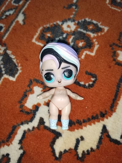 A close-up, top-down shot captures a small, plastic doll positioned on a vibrantly patterned carpet. The doll has large, striking blue eyes with black eyeliner and light-colored eyelashes. Its hair is styled in a vibrant, multi-colored sweep, featuring streaks of black, white, light purple, and pastel blue. The doll's skin is a pale peach tone, and it has delicate facial features. On its left arm, there are three small, white star tattoos, and a faint lightning bolt tattoo is visible on its chest. The doll is wearing light blue socks adorned with a repeating white pattern of short, diagonal lines. Its body is otherwise unclothed. The carpet beneath the doll features a bold, geometric design in shades of orange, black, and cream, with a slightly fuzzy texture. The lighting suggests an indoor setting, and no specific time of day or weather conditions are indicated. The image was taken in Aksay, Kazakhstan.