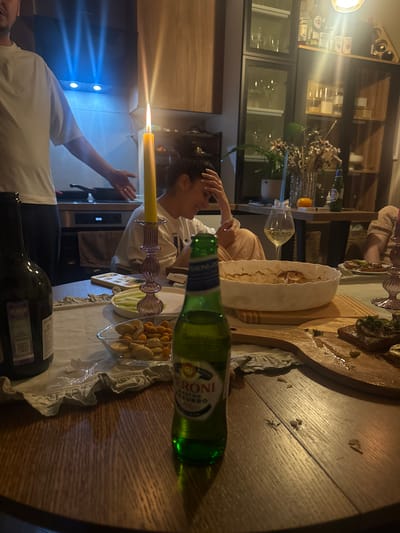 The image depicts an indoor dining scene. Two individuals are visible: one gesturing with an outstretched arm and a second, laughing, with a hand on their head. A table dominates the foreground. On it are an assortment of items including a lit candle in a glass holder, an unopened bottle of Peroni beer, a wooden board with food, a bowl, glassware, a dish with snacks, and a bottle of wine. The background features a kitchen area with cabinets, appliances, and decorative elements such as floral arrangements.
The information provided suggests the location is Razvilka, Russia, but the image itself offers no means to confirm this.