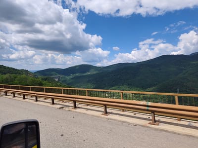 This is a wide-angle, eye-level shot taken from a vehicle, likely a car or truck, moving along a road. The foreground is dominated by the asphalt road surface and a rusty brown metal guardrail that runs along the side of the road, parallel to the camera's movement. The guardrail is supported by metal posts embedded in a concrete barrier. In the lower-left corner of the frame, a side-view mirror of the vehicle is visible, reflecting a blurred image of what appears to be a person.
The middle ground of the image features a lush, green, mountainous landscape. The slopes of the mountains are densely covered with trees. In the distance, a large, concrete bridge or dam structure is visible, spanning across a valley. Power lines can also be seen on one of the distant hills.
The sky above is a vibrant blue, dotted with fluffy white cumulus clouds. The lighting suggests it is daytime, likely late morning or early afternoon, with the sun casting shadows on the mountains. The overall scene depicts a scenic drive through a hilly, forested region, possibly in Bulgaria, given the location context. There is no visible text in the image.