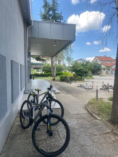 The image shows an outdoor scene with several bicycles parked on a paved area. In the foreground, two mountain bikes are prominently displayed, leaning against each other. Their wheels and frames are clearly visible. Further back, additional bicycles are parked on the pavement, some of which appear to be leaning against a bike rack.
The setting appears to be a residential or public area, with buildings and trees visible in the background. There are houses with red tiled roofs and lush green trees and bushes. The ground is paved with stone tiles. The sky is blue with fluffy white clouds, suggesting it is daytime and the weather is pleasant. A large tree branch partially frames the top of the image.
One of the mountain bikes has a yellow and blue accent on its frame and the word "GALAXY" is visible on the tire. There is also some text on a white bag in the background, which is partially obscured and illegible.
The overall impression is a peaceful and ordinary day, with bicycles parked and ready for use. There are no people visible in the frame, giving a sense of quietness. The location context suggests this scene is in Ovoshtnik, Bulgaria.