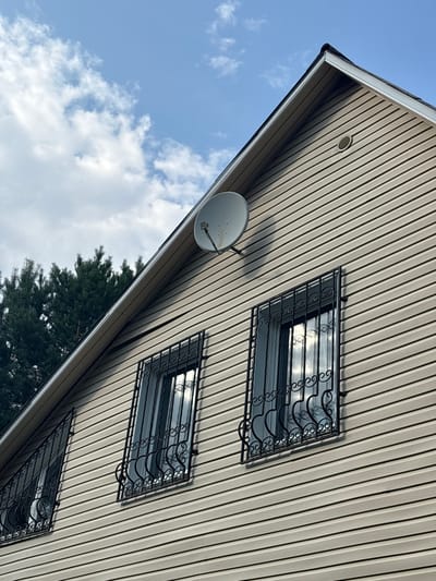 The image shows a close-up, low-angle view of a house with beige vinyl siding under a cloudy blue sky. A dark green evergreen tree line is visible on the left side of the frame, behind the house. The house has a steeply pitched roof. A white satellite dish is mounted on the siding near the apex of the roof. Three rectangular windows with decorative black metal bars are visible on the side of the house. The windows reflect the sky and clouds. A circular ventilation grate is also visible on the siding. The scene appears to be in a residential area, possibly in the city of Lyady, Russia, based on the prompt's location information.