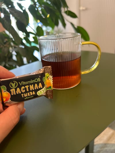 An individual is holding a wrapped candy in their left hand, positioned in the foreground of the image. The candy wrapper is black with colorful illustrations of fruit and text. Behind the candy, a clear glass mug filled with a dark amber liquid, likely tea, is visible. The mug has a ribbed texture and a golden handle. The setting appears to be indoors, with a dark green table surface in the foreground. In the background, blurred green foliage suggests the presence of plants, and a light-colored vertical surface, possibly a wall or door, is also visible. The scene is well-lit, possibly by natural light. No specific location in Medvedevo, Russia, is indicated by any visible elements in the image.