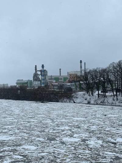 The image depicts an overcast winter scene in Мастоўскі сельскі Савет, Belarus, showing an industrial complex across a partially frozen river. The foreground features a wide body of dark grey water, largely covered by a dense accumulation of irregularly shaped, flat white and light grey ice floes. No individuals or vessels are present on the water.

The midground consists of the snow-covered riverbanks. On the left, a dense line of bare, dark deciduous trees and low-lying brush, also dusted with snow, extends along the bank. On the right, a higher, snow-covered embankment supports a row of widely spaced, tall, bare deciduous trees. A dark, low structure is partially visible in the water near the right bank.

In the background, an industrial facility spans the horizon. It comprises multiple light-colored buildings with green and beige facades. Prominent structures include several smokestacks: two large, striped light blue and white stacks are centrally located, surrounded by complex piping and scaffolding. To their right, several thinner, dark grey smokestacks of varying heights rise above other industrial buildings. The sky above is a uniform, pale grey, indicating an overcast day. No human activity or movement is discernible within the industrial complex.