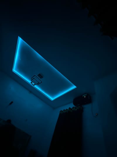 A dim room, illuminated by a rectangular recessed ceiling light that emits a vibrant blue glow, highlights an unusual overhead fixture that looks like a printer. Above and right is a dark projector, partially obscured. To the right, in the same area, an equally dark curtain hangs. Other features, like furniture and additional decor, are indistinguishable due to the poor lighting conditions.