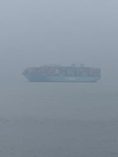 The image shows a cargo ship navigating the waters off the coast of Southend-on-Sea, United Kingdom. The ship is heavily laden with shipping containers. The atmospheric conditions are foggy and overcast, significantly obscuring the visibility of the ship and the surrounding seascape. The water appears calm with a gray hue, blending with the sky and creating a low-contrast scene.