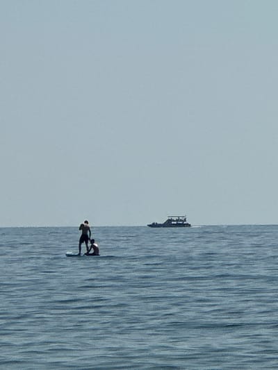 Two people are on a paddleboard in the sea near Đurovići, Montenegro. The person in front is standing and holding a paddle, while the person behind is kneeling. A boat is visible in the distance. The water is calm with small waves, and the sky is clear and pale blue. The scene appears to be during daylight.