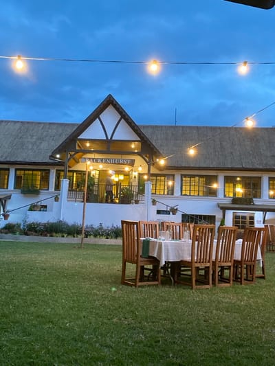 This photograph, taken in Riara Ridge, Kenya, captures an outdoor evening scene featuring a white, two-story building with a distinctive gabled entrance. The building, identified by a sign reading "BRACKENHURST" above its main entrance, has numerous dark-framed windows that reflect the interior lighting. Its architectural style incorporates dark timber framing on the gable, reminiscent of half-timbered designs.
Across the deep blue, twilight sky and adorning the building's facade, multiple strands of illuminated string lights cast a warm glow over the scene. In the foreground and extending to the right, a well-maintained green lawn is visible. Arranged on this lawn are several outdoor dining tables, each set with a white tablecloth, glasses, and what appear to be green placemats or napkins. Matching wooden chairs are placed around these tables, suggesting a setting prepared for an outdoor event or dining experience. A blurry figure is vaguely discernible inside the building near the entrance. Potted plants hang from various points on the building and from a wooden pole on the lawn. The overall impression is one of a serene and inviting ambiance at dusk or early evening.