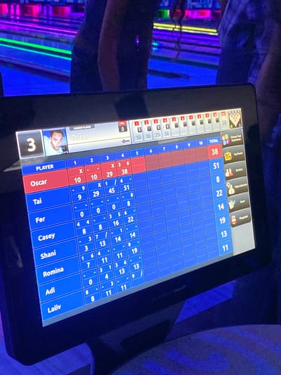 The image shows a close-up of a bowling alley's scoring touchscreen. The screen displays a bowling game in progress, with several players' names and scores listed. The current player is identified as "Oscar." The scoreboard shows frames 1 through 10, with scores for each frame and a total score for each player. Names visible on the scoreboard include Oscar, Tai, Fer, Casey, Shani, Romina, Adi, and Laliv. The background of the image is blurred and shows the colorful lights of the bowling lanes, typical of a bowling alley environment. The scene is dimly lit, with the primary illumination coming from the touchscreen display and the bowling alley lights. The image does not provide any specific location information within Waynesboro, United States of America, but it is a typical bowling alley scene.