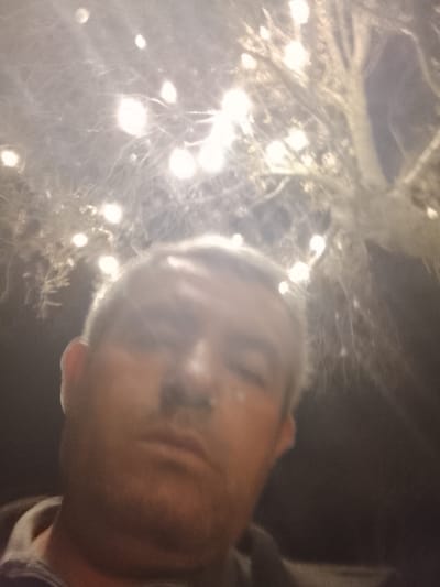 The photograph shows a man with fair skin and short, graying hair in a selfie-style image. His face is the primary subject, filling much of the frame. Above and behind him, there appears to be a tree or shrubbery illuminated with numerous small, bright lights. The background is dark, suggesting it is nighttime. There is no indication of location in the city of Sabir Rakhimov, Uzbekistan from the image itself.