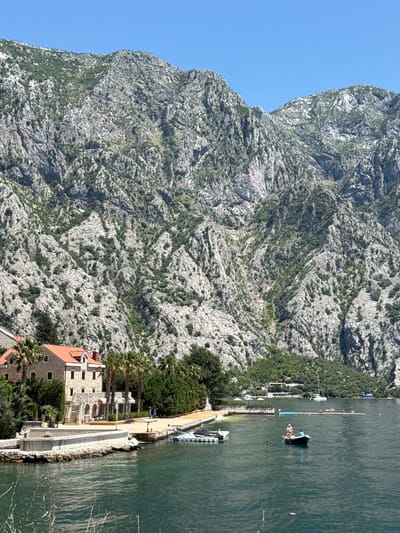 This image depicts a sunny coastal landscape in Donji Orahovac, Montenegro, characterized by a dramatic mountainous backdrop, a calm bay, and waterfront structures.

The upper two-thirds of the image are dominated by towering, steep, light-grey limestone mountains, heavily fissured and sparsely covered with green vegetation, including shrubs and small trees clinging to the rock face. The sky above is a clear, bright blue.

In the lower third, a deep green-blue body of water occupies the foreground and mid-ground. On the immediate left foreground, a prominent multi-story building with a light stone facade and a reddish-orange tiled roof stands by the water's edge. This building features multiple windows, an arched ground level, and a visible chimney. A stone terrace with stairs and arches extends from the building towards the water, flanked by several tall palm trees and other dense green foliage. A concrete pier or wall extends from this property into the water, with a small white boat moored alongside it.

In the mid-ground water, a dark-colored rowboat or small motorboat is visible, carrying two individuals. One person, wearing a bright orange top or life vest, is seated facing forward, while the other is rowing, facing away from the viewer. Further across the bay, a long, blue floating platform is present, with several people on and around it, suggesting a swimming area. Beyond this, a small sandy beach with sun umbrellas is discernible on the far shore, with additional buildings nestled among trees at the base of the mountains. Several larger boats, including sailboats, are anchored further out in the bay.

Sparse, out-of-focus green reeds or grass blades are visible in the extreme lower foreground. The scene is brightly lit by natural daylight, casting clear but not overly harsh shadows.