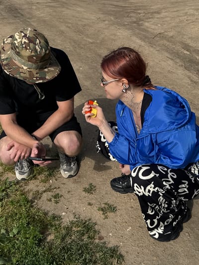 Two individuals are crouched down on a dirt path, a grassy area is visible to the left. The individual on the left wears a camouflage bucket hat, a black t-shirt, and shorts, and is holding a smartphone. The individual on the right has red hair, wears a blue jacket and black pants with white lettering, and is holding a small, colorful object, possibly a toy. The background appears to be an outdoor, possibly urban or semi-urban environment. There is no specific visual information in the image to confirm or deny the location as Desnogorsk, Russia.