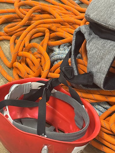 The scene, located in Tayshet, Russia, displays an arrangement of climbing or safety equipment on a light-brown wooden or laminate floor. Multiple coils of orange kernmantle rope, featuring integrated blue tracer strands, occupy the central and upper portions of the frame. A segment of blue and white braided rope is partially visible beneath the orange ropes. In the lower left quadrant, an inverted red hard-shell helmet is present, revealing its internal harness system comprising black webbing straps, grey padding, and a white plastic buckle. A grey, textured fabric bag or backpack component, displaying a black zipper and dark mesh material, rests on the ropes in the upper right. No individuals are depicted.