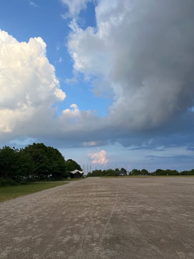 The image presents a wide outdoor landscape under a dramatic sky. The foreground and midground are dominated by a vast, flat expanse of light brown gravel or dirt, showing subtle tracks on its surface. Along the horizon, a dense line of dark green trees stretches across the width of the frame. Interspersed among and just above the trees are several slender white mast-like structures, suggesting the proximity of a marina or harbor. A few dark-colored buildings are also partially visible behind the foliage. On the left side of the tree line, a large white, tent-like canopy structure can be seen.
The sky above is a striking blend of elements. On the left and upper parts, patches of bright blue sky are visible, contrasting sharply with large, heavy dark grey storm clouds that cover much of the upper portion of the sky. Fluffy white cumulus clouds are also present, with some displaying a subtle pinkish-orange hue, particularly towards the right side of the sky, indicating either a sunset/sunrise or a specific atmospheric condition during late afternoon or early evening. The overall weather appears dynamic, with clear skies battling against an impending or receding storm.
No people or visible text are present in the scene, which is static. The location is Aarhus, Denmark, with the mast structures providing a strong cue of its coastal nature.