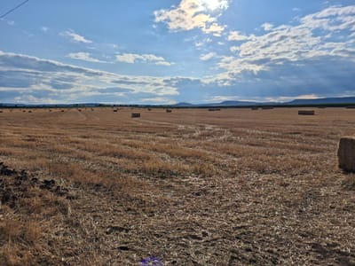 An outdoor shot captured in Goran, Bulgaria, features a vast, dry field dotted with numerous square hay bales. The field is covered in harvested straw, indicating recent agricultural activity. The sky above is a mix of blue and white clouds, with sunlight breaking through in some areas, casting light beams downwards. In the distance, a range of low hills forms the horizon. The overall scene depicts a rural landscape during what appears to be the aftermath of a harvest.