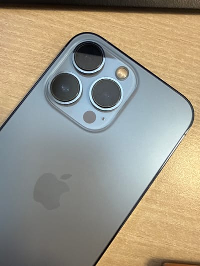 A close-up, overhead shot focuses on the back of a silver iPhone 13 Pro, angled slightly to the right. The device rests on a light brown wooden surface with a visible grain. The prominent camera module, featuring three circular lenses and a flash, occupies the upper left portion of the phone. The Apple logo is subtly visible in the lower left quadrant of the phone's back. The image does not provide any information about the city of Gülnar, Turkey, or any individuals present.
