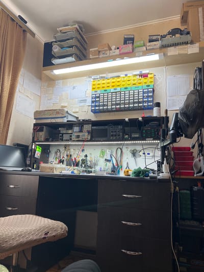 This media file captures an indoor scene, rotated 90 degrees clockwise, depicting a cluttered yet organized electronics workshop or repair bench. No people are visible.

The scene is dominated by a long workbench illuminated by two white fluorescent lights. On the left side of the bench, various electronic components are neatly stored in multi-drawer organizers (yellow, blue, green). Above these, shelves hold stacked items, including what appear to be server-like units or heat sinks. The wall behind the bench is densely covered with numerous handwritten notes, diagrams, and technical information, primarily in Cyrillic script.

The central area of the workbench is equipped with an array of electronic test and repair equipment, including a LUKEX 702 soldering station, power supplies, a multimeter, and other devices, all connected with various wires and cables. Hand tools such as scissors, pliers, screwdrivers, and brushes are arranged on the white surface. Boxes and containers with brand names like "NEVA LUX," "VAILLANT," "PHILIPS," "HMER," "Thermex," "SAMSUNG," and "won Digital Multi" are scattered around. A black trash bag is visible near the top left, and a monitor screen is partially visible at the bottom.

To the right, a dark wooden cabinet with three distinctive metallic handles suggests storage. A beige, textured chair is partly visible in the foreground, indicating a user's presence.

The presence of specialized tools, components, and technical notes strongly suggests an active workspace for electronics repair, development, or hobbyist activity. The lighting implies work is either ongoing or frequent. The visible text, including brand names and Cyrillic notes, provides specific product and technical context.