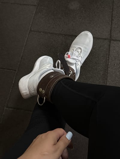 In Nazran, Russia, a person is sitting on a dark gym floor. They are wearing white athletic shoes and black leggings. A brown leather ankle strap with a metal ring is attached to their left ankle, just above the shoe. Their right hand, with light blue nail polish, rests on their leg. The gym floor is made of dark, interlocking rubber tiles.