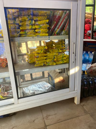 The image depicts a retail display cabinet situated in Okura, Nigeria, observed from a slight angle. The white-framed glass cabinet occupies the majority of the foreground and center, showcasing various packaged goods. The top and middle shelves are filled with numerous stacks of yellow-packaged instant noodles. The bottom shelf, visible through the glass, contains a clear plastic bag with contents, a cardboard box, and, on the far left, multiple brown-packaged items labeled "JUMBO."
To the immediate right of the cabinet, several plastic bottles of water with blue caps are stacked, alongside multiple dark-colored plastic bottles labeled "V-COOL" arranged on the tiled floor. The floor itself is light-colored tile, showing some dirt and wear.
Through the reflective glass and the open background area to the right, an outdoor environment is visible. This includes a blurred individual partially obscured by red fabric, a metal grid structure, and distant vegetation. A distinct beam of sunlight illuminates the right side of the cabinet and a portion of the floor. No specific actions or interactions are discernible from the individuals present in the background.