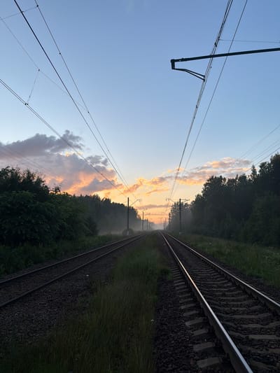 The image depicts a railway line at dawn or dusk in the city of Orekhovo, Russia. The sky is a soft gradient of blue and pink, with illuminated clouds in the distance suggesting the sun is either rising or setting. Overhead, a complex network of electrical wires for the trains crisscrosses the frame. On either side of the tracks, dense foliage and trees form a natural border, with some areas showing a slight haze or fog close to the ground. The railway tracks themselves, composed of gravel and sleepers, stretch into the distance, converging towards a vanishing point. There are no people or animals visible in the scene. The overall impression is one of quiet, solitary beauty in an industrial landscape.