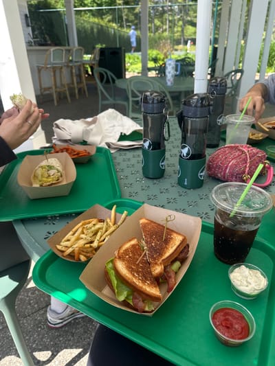This is a close-up, eye-level, outdoor shot of a meal on green trays at what appears to be a sports venue, possibly a tennis club, in Harbor Springs, Michigan. In the foreground, a green tray holds a toasted sandwich cut in half, french fries, a cup of soda with ice and a green straw, a small container of ketchup, and a small container of dip. A second green tray, partially visible, holds a wrap and a side of what looks like sweet potato fries. Three dark water bottles with green sleeves sit on the table. In the background, a person is visible on a tennis court, and a building with bar stools and tables suggests an outdoor seating area. The overall impression is that of a casual meal enjoyed during a sporting event.