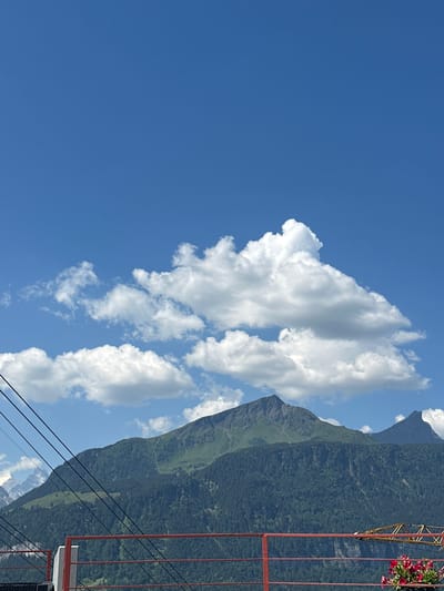 In Hasliberg Reuti, Switzerland, a mountain landscape unfolds under a clear blue sky dotted with white cumulus clouds. The foreground features a red railing, with some cables or wires extending diagonally across the scene from the left. Beyond the railing, a dense, dark green forest covers the slopes of a large mountain. To the right of this mountain, another, more pointed mountain peak is visible. The summit of the larger mountain displays patches of green, suggesting grassy areas amidst the darker forest. The image captures a serene, natural environment.