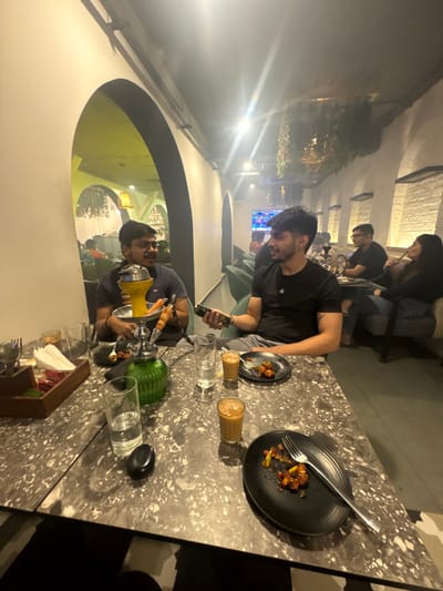 A group of people sit at a table. One man holds a phone, near food and drinks. Another has a hookah pipe. Black plates with food remnants and a fork sit on the patterned table. Glasses of water, small teacups, and hookah components are also present. The setting appears to be an outdoor patio or restaurant, possibly with television screens in the background. Arched architecture is visible in the background.
