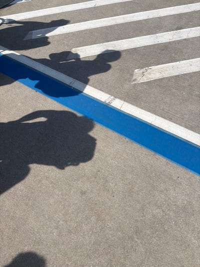 The image shows shadows of people cast on a paved surface, likely a sidewalk or parking lot, marked with white and blue lines. The shadows suggest at least three individuals, with one shadow appearing larger and more detailed, possibly a person holding a phone. Another shadow is elongated, and two smaller shadows are also visible. The lines on the pavement are painted, with white lines indicating parking or directional markings and a prominent blue line. The surface is concrete, with some visible cracks and wear. The bright sunlight indicates it is daytime, and the clear shadows suggest fair weather. The scene is an outdoor urban or semi-urban environment. There are no discernible emotional cues or specific event indications, other than the presence of people. No visible text is present in the image. The location context is Balice, Poland.