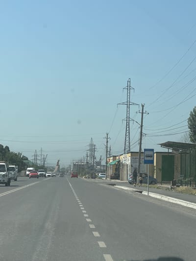 The image depicts a street scene in the city of Ынтымак, Kyrgyzstan, on a clear, sunny day. The asphalt road is divided by a dashed white line, with vehicles traveling in both directions. On the left side of the road, several cars and trucks are visible, including a white truck in the foreground. On the right side, there are small buildings with storefronts and a bus stop sign. Power lines crisscross the sky, supported by tall metal towers and wooden poles. In the distance, a large statue of a person is visible. The environment appears to be a commercial or mixed-use area on the outskirts of the city, with a modest urban landscape. Two individuals, an adult and a child, are walking on the sidewalk near the bus stop.