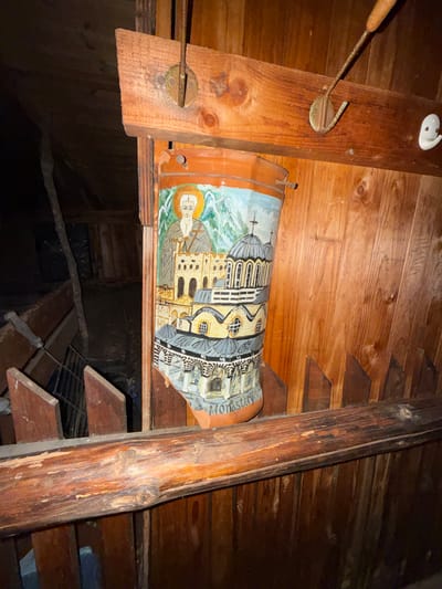 An eye-level interior shot shows a wooden structure, possibly an attic or crawl space, in Rila, Bulgaria. A painted clay beer stein rests horizontally on a wooden beam. The stein features an image of a religious figure and architectural buildings labeled "Monasteri." The area beneath the stein is dark and cluttered. Tools are attached to the wood, including a small hand-held garden rake. Wooden stairs ascend to the right of the scene.