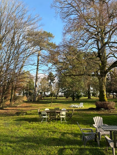 This media file depicts an outdoor garden or park setting, likely on a sunny day. The scene is dominated by a clear, bright blue sky visible through the canopy of numerous trees.

The foreground and midground feature a variety of trees. Many are mature deciduous trees with bare branches, some covered in moss or lichen, indicating late winter or early spring. Interspersed among these are evergreen trees with green foliage. The ground is covered in lush green grass, with patches of sunlight and shadow cast by the trees.

Scattered across the grassy area are several sets of outdoor patio furniture. These include tables and chairs, arranged in small clusters. Some chairs are light-colored, possibly plastic, while other furniture pieces appear darker, possibly made of wood or metal. Some chairs are stacked or casually placed.

No people are visible in the image, suggesting a quiet moment before or after an event, or simply a tranquil leisure area. There is no specific activity taking place. The time of day is daytime, evidenced by the bright light and clear shadows. The weather is sunny with a cloudless sky. The location context of Étréchy, France, aligns with a garden or park setting suitable for outdoor relaxation. There is no visible text.