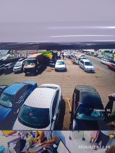 The image is a CCTV still that appears to be taken in a parking lot. The timestamp in the upper left corner reads "2026 Mon 14:49:32". The scene shows several cars parked, including a dark minivan, a white sedan, and a blue sedan. In the background, there are buildings that suggest an urban or commercial environment, with some trees visible. A red object, possibly a car or a large piece of equipment, is also present in the distance. The ground is paved. The overall lighting suggests it is daytime. The bottom portion of the image is a split screen, showing a different scene with people, possibly shoppers, and a timestamp "2026/6/29 14:49" in the lower right corner.
While the image is a CCTV still, there is no specific visual information within the image that allows for the definitive identification of the location as Ak-Suu, Kyrgyzstan. The general appearance of the buildings and vehicles is not uniquely characteristic of this specific city or region.