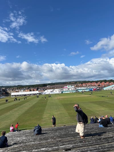 An outdoor photograph captured on a bright, sunny day displays the North Marine Road Ground, a cricket stadium in Scarborough, United Kingdom. The foreground features worn, tiered wooden bleachers, occupied by approximately eight individuals. A male subject, positioned centrally, stands wearing a dark long-sleeved top and light-colored shorts, his right hand raised to shield his eyes. Another male in a black t-shirt and shorts stands further down the seating. Other seated spectators are visible. The midground is dominated by a meticulously maintained green cricket pitch, marked with distinct mowing stripes and containing multiple cricket wickets. Approximately 20 to 30 individuals, presumed to be players or ground staff dressed in dark athletic wear, are scattered across the field, engaged in activities such as walking, standing in small groups, or light warm-ups. The background reveals a series of spectator stands, including temporary white marquee structures, leading up to a residential area of terraced houses with red roofs and a tree-covered hillside under a partially cloudy blue sky.