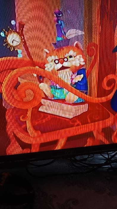 A digital screen, displaying an animated scene, is captured, exhibiting prominent vertical scan lines. The animated content features an anthropomorphic orange-and-white character, resembling a feline, wearing round spectacles and a blue garment. This character is seated at an ornate, orange-brown scrollwork desk or chair, interacting with an open book displaying multicolored pages. A small, purple, conical figure is positioned on the character's head. To the left of the main character, a stylized golden clock with a sunburst-like frame and a white face is visible. Below the illuminated screen, the dark physical bezel of a monitor or television is partially visible, showing a faint white logo on the lower right. A tangle of black electrical cables rests on a dark surface directly beneath the screen. The background behind the cables is a dimly lit wall or surface patterned with dark, swirling motifs. This observation takes place in the city of Kasansay, Uzbekistan.