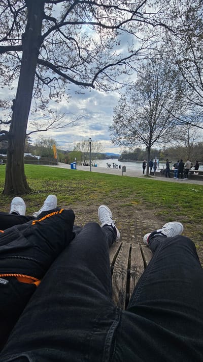 A casual photograph shows a group of people sitting on a bench near the Rhine river in Rheinfelden, Switzerland, during the day. The image is taken from a first-person perspective, showing the legs of three people sitting on a wooden bench. One person wears dark denim jeans and Adidas sneakers, while another wears dark pants with orange trim and white sneakers. A third person has their legs in front, wearing dark jeans and white sneakers. In the background, a tree with bare branches is visible, along with the Rhine river and a grassy area. There are some other people standing or sitting on benches nearby, enjoying the riverside.