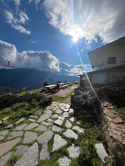 The image depicts an outdoor seating area of a mountain hut or lodge, likely situated at a high elevation, offering a panoramic view of a vast mountain range and valley. In the foreground, a paved pathway made of irregular flagstones, interspersed with grass, leads towards the lodge. To the left of the pathway, a low stone wall borders a grassy slope. A flagpole with the Bulgarian flag is visible on the left side of the image.
The lodge itself, constructed with stone and painted white, stands on the right side of the frame. It features a prominent sloped roof and a window. A noticeboard is affixed to the stone wall of the lodge. In front of the lodge, several people are seated at picnic tables. The sun is shining brightly in the sky, creating lens flare and illuminating the scene. The sky is a vibrant blue with dramatic white clouds. The background shows layered mountains fading into the distance, enveloped in shadow, suggesting the setting sun or a cloudy day over the peaks. The overall impression is one of a scenic and tranquil outdoor gathering place in a mountainous region. The location is identified as Rozino, Bulgaria.