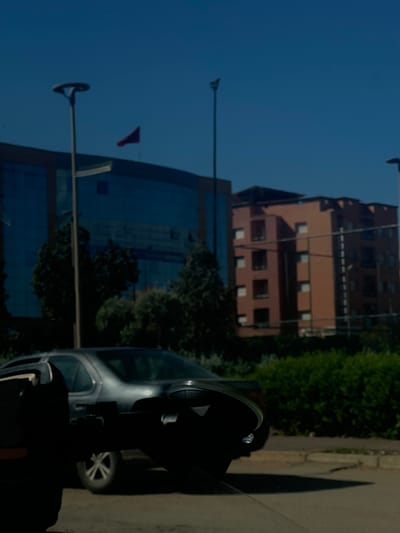 The image presents an urban street scene in Beni Mellal, Morocco, captured during daylight hours under a clear blue sky. The immediate foreground, viewed from a vehicle's interior, shows a dark, indistinct object (possibly a device mount or part of a rearview mirror) in the lower center, partially obscuring a dark gray sedan positioned ahead on a paved road. Beyond the road, a strip of dense green vegetation, primarily bushes and trees, is visible. Two tall streetlights stand prominently, one in the mid-left and another further back on the right. In the background, two primary structures are apparent: a modern building with reflective blue glass facades on the left, featuring a red flag flying from a flagpole on its roof, and a multi-story, terracotta-colored building with numerous rectangular windows on the right. No individuals are visible in the scene.